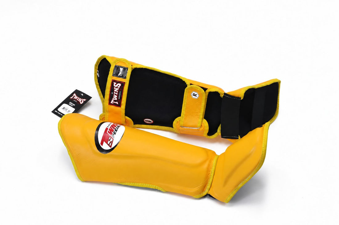 Angled view of yellow Twins Special SGL-10 shin guards showing contoured shin and instep padding, secure hook-and-loop closures, and padded foot support for Muay Thai and kickboxing on a white background