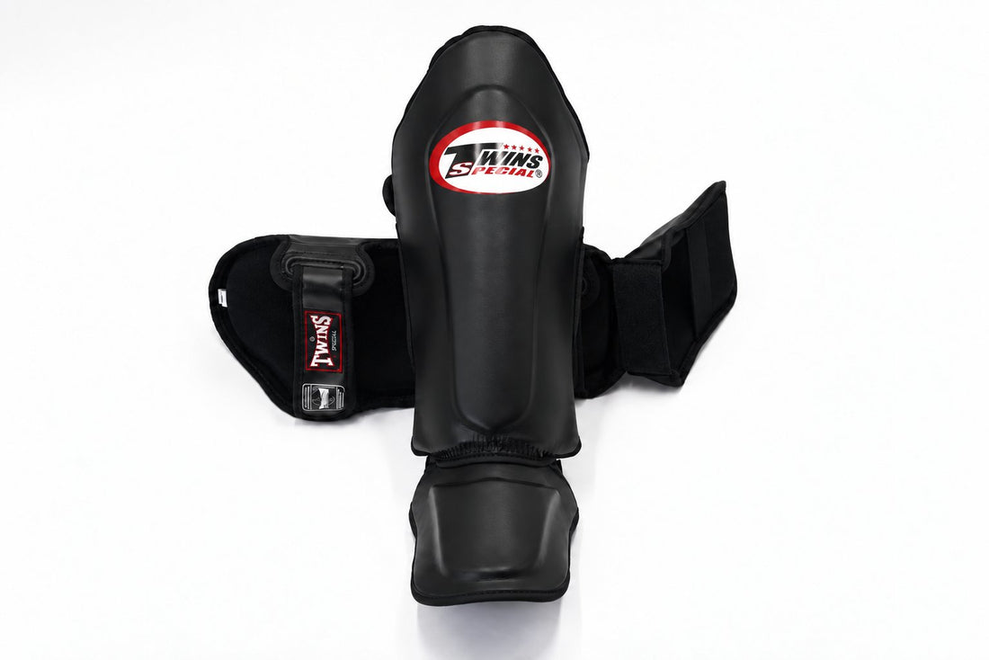 Front view of black Twins Special SGL-10 shin guards showing full-length shin and instep padding, secure hook-and-loop straps, and padded foot support for Muay Thai and kickboxing on a white background