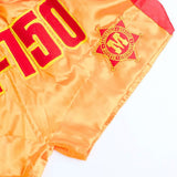 Close-up of gold and red ThaiSmai M-150 Golden Era Muay Thai shorts showing satin fabric, embroidered lettering, crest patch and side seam on a white background