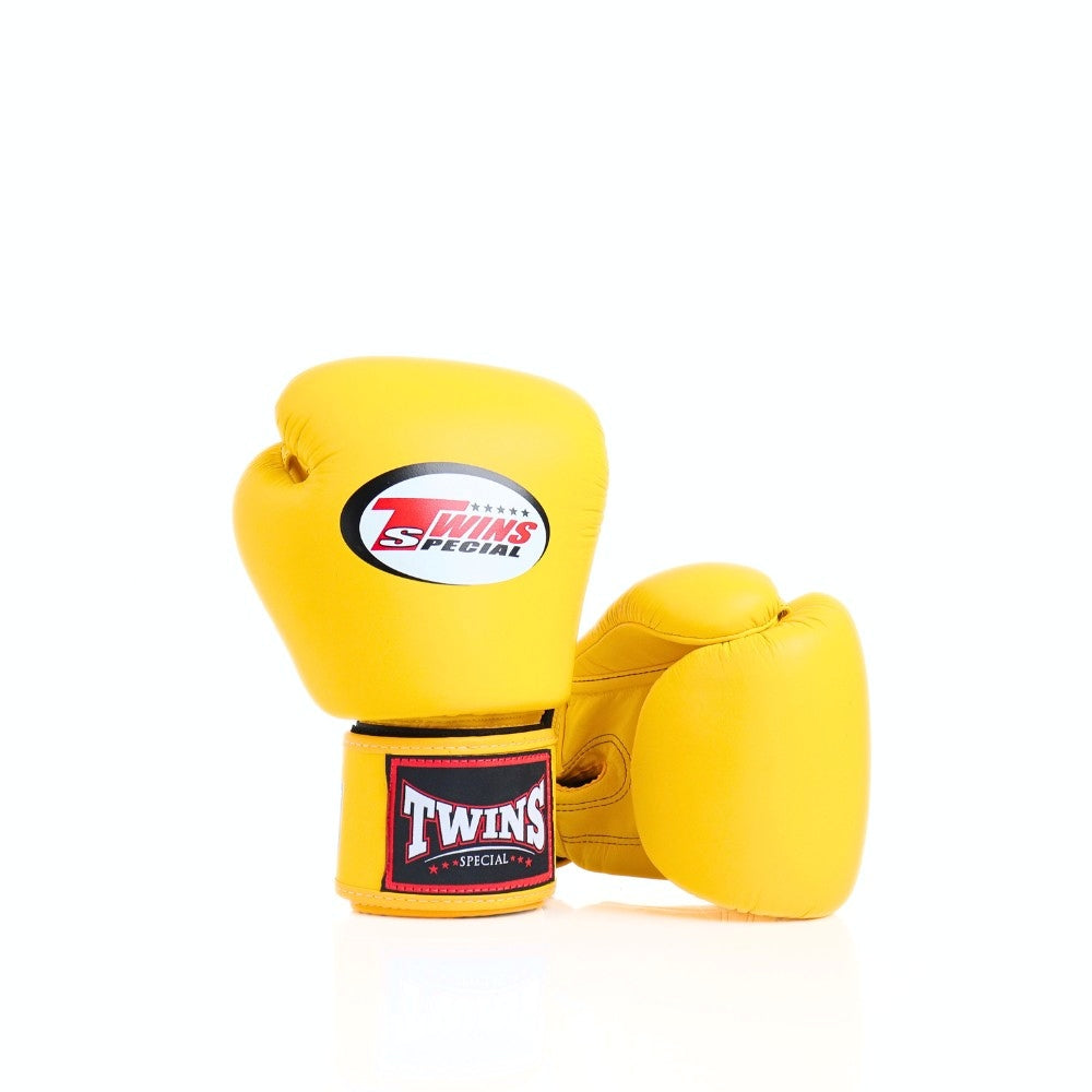 Pair of yellow Twins Special boxing gloves with one glove upright showing the logo and wrist patch while the other lies behind on a white background