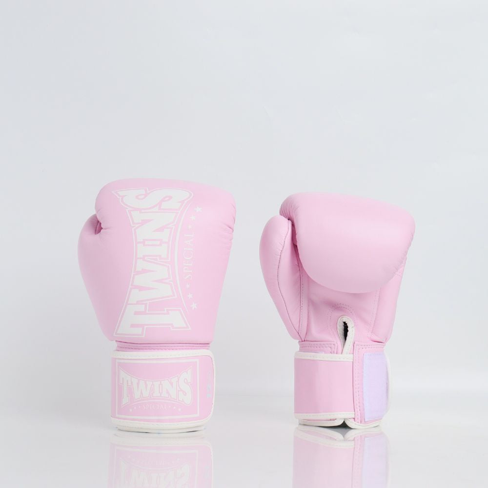 Pair of light pink Twins Special boxing gloves showing front branding and rear hook-and-loop wrist closures on a reflective white surface